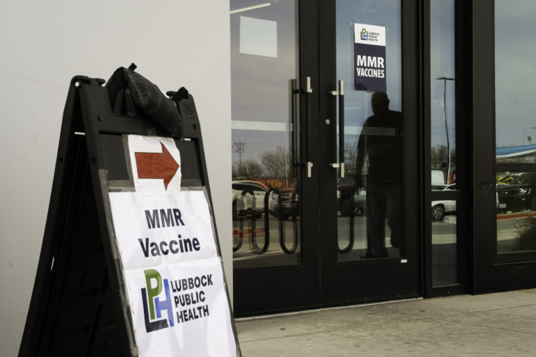 Whooping Cough & Measles outbreak: Sign on sidewalk outside double glass doors with red arrow ant black text on white "MMR vaccine, Lubbock Public health""