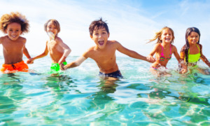 Vitamin D deficiency in children: Mixed ethnic group of 5 young children in swimsuits hold hands and run in clear turquoise water along beach