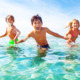 Vitamin D deficiency in children: Mixed ethnic group of 5 young children in swimsuits hold hands and run in clear turquoise water along beach