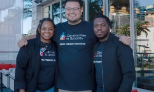 CIS and Whataburger Partnership: Three adults wearing black t-shirts with the CIS logo and Communities in Schools text stand at a double glass door entrance of modern building