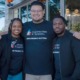 CIS and Whataburger Partnership: Three adults wearing black t-shirts with the CIS logo and Communities in Schools text stand at a double glass door entrance of modern building