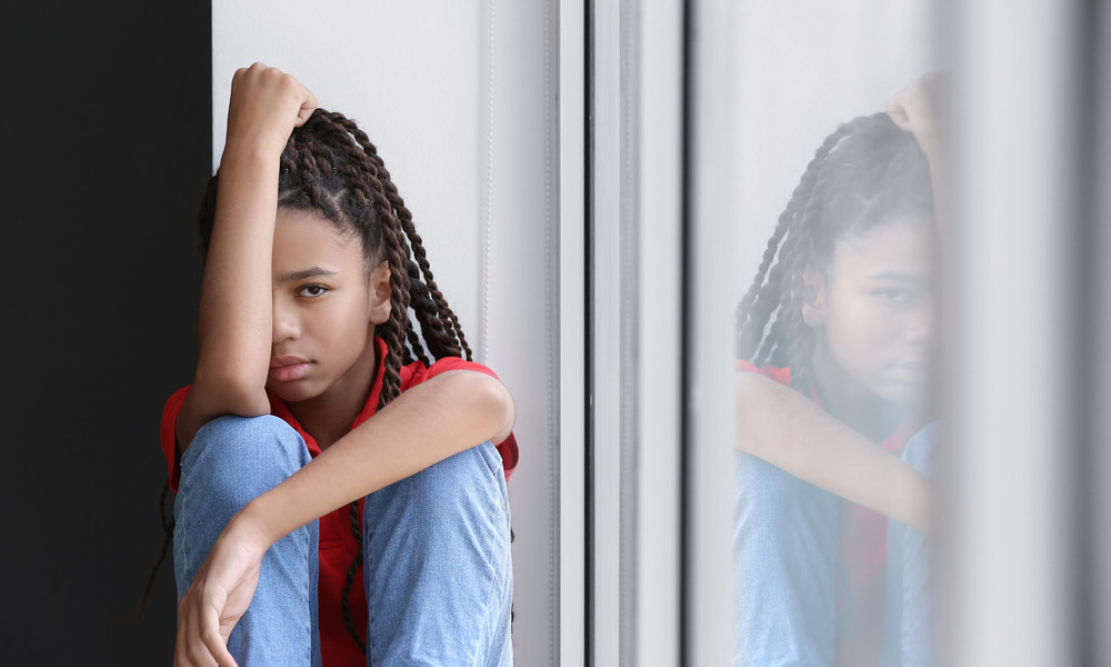 Children recovery after disaster: Sad Black preteen girl with long braids in red shirt and blue jeans sitting on windowsill.