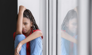 Children recovery after disaster: Sad Black preteen girl with long braids in red shirt and blue jeans sitting on windowsill.