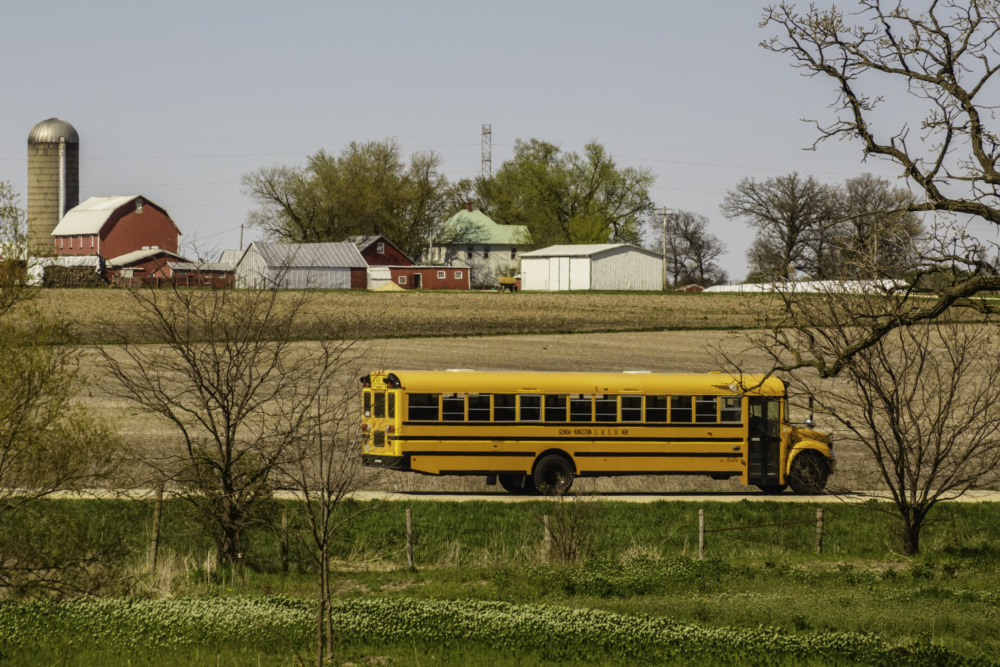 What rural students need: Yellow school bus driving alone on road between country pastures with farm buildings in far background