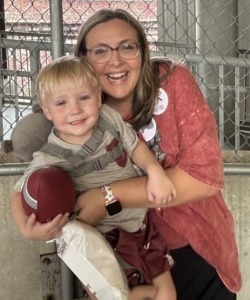 What rural students need: Headshot blonde woman with long in pink cardigan and white blouse crouched on the floor holding a blonde boy toddler