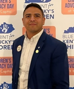 What rural students need: Headshot man with dark hair in dark suit with white shirt staning in fron of backdrop with pink and blue figures and text