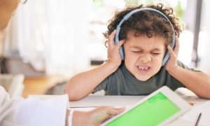 Autism Kennedy versus CDC: Young boy wearing earphones sits at table face scrunched up, eyes closed, and holding hands over ears