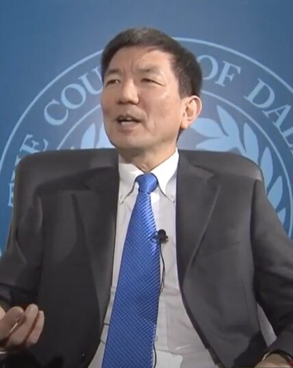 Whooping Cough & Measles outbreak: Asian man in charcoal suit, white shirt and blue tie speaking in front of blue Dallas County shield.
