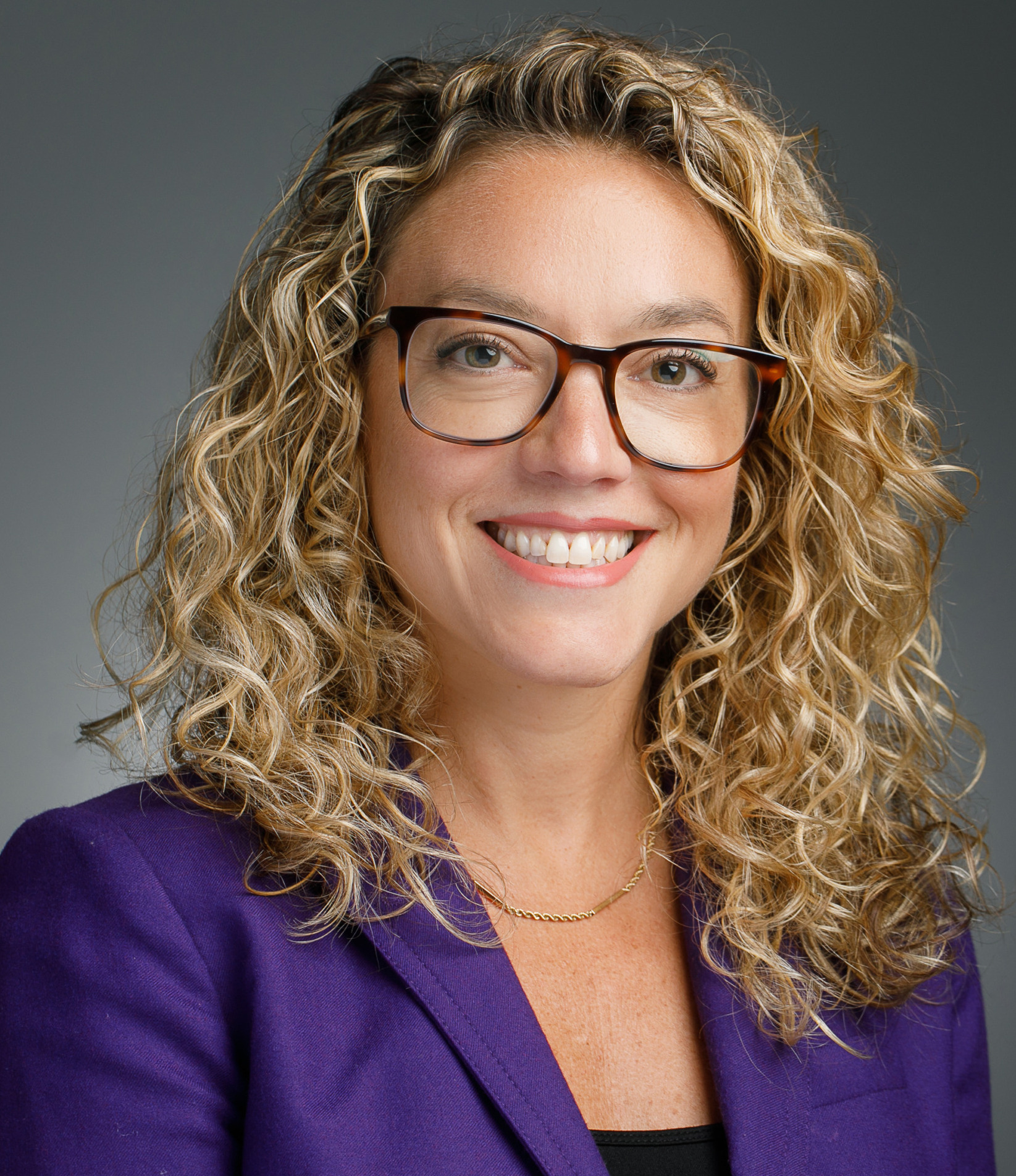 Legal counseling for behavior issues: Headshot woman with long, curly, blonde hair and dark framed glasses in purple suit with dark top against gray background