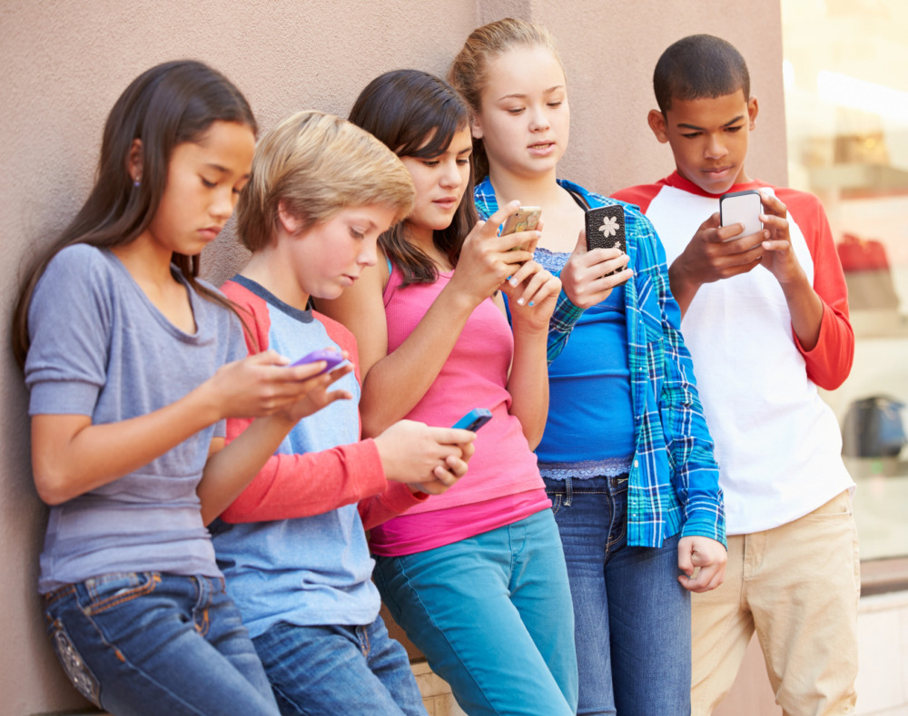 Cell phones in middle school: 5 middle schhol age kids of mixed ethnicity and gender stand leaning agaist a wall all holding cell hones and looking at screens