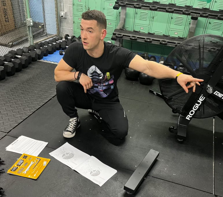 School cell phone ban: Man with blonde hair in black t-shirt and pants squats on black gym floor matts surrounded by free-weight lifting equipment with left arm pointing to his left.
