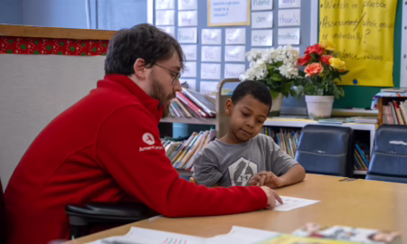 Americorps funding cuts: Man with brown hair, beard and glasses, in red Americorps sweatshirt sits at student desk in classroom helping young boy with class work