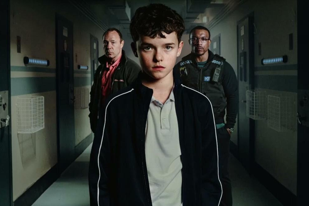 Adolescence Netflix series: young teen with short dark hair, light shirt ad dark sweat jacket tands in foreground or dimly lit room with two adult men -- one white , one black --several steps behind him on either side