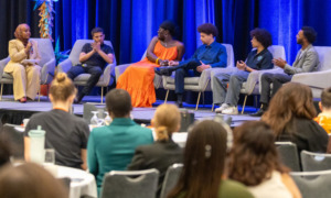 Youth leadership:Panel of 6 people seated on stage in light grey chairs in front of royal blue curtain with backs of heads of audience in foreground.