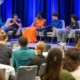 Youth leadership:Panel of 6 people seated on stage in light grey chairs in front of royal blue curtain with backs of heads of audience in foreground.