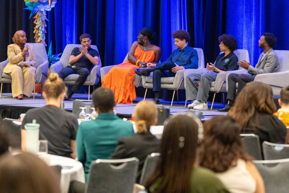 Youth leadership:Panel of 6 people seated on stage in light grey chairs in front of royal blue curtain with backs of heads of audience in foreground.