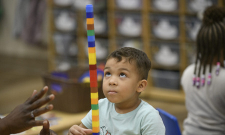 Transitional Kindergarten: Young, dark-haired boy sits at table staring up to top of staked pile of multi-colored blocks