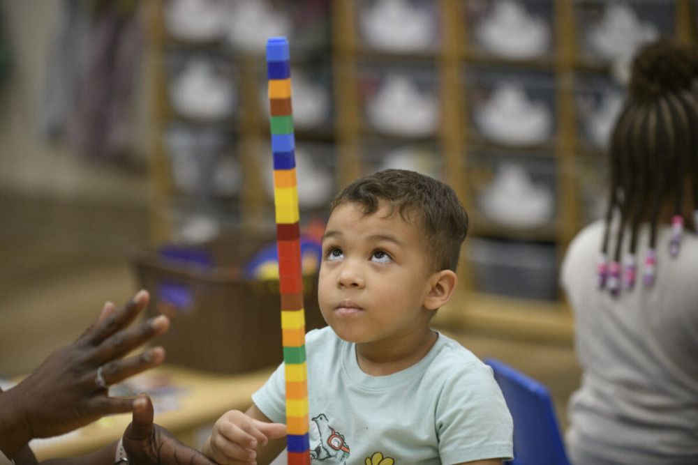 Transitional Kindergarten: Young, dark-haired boy sits at table staring up to top of staked pile of multi-colored blocks