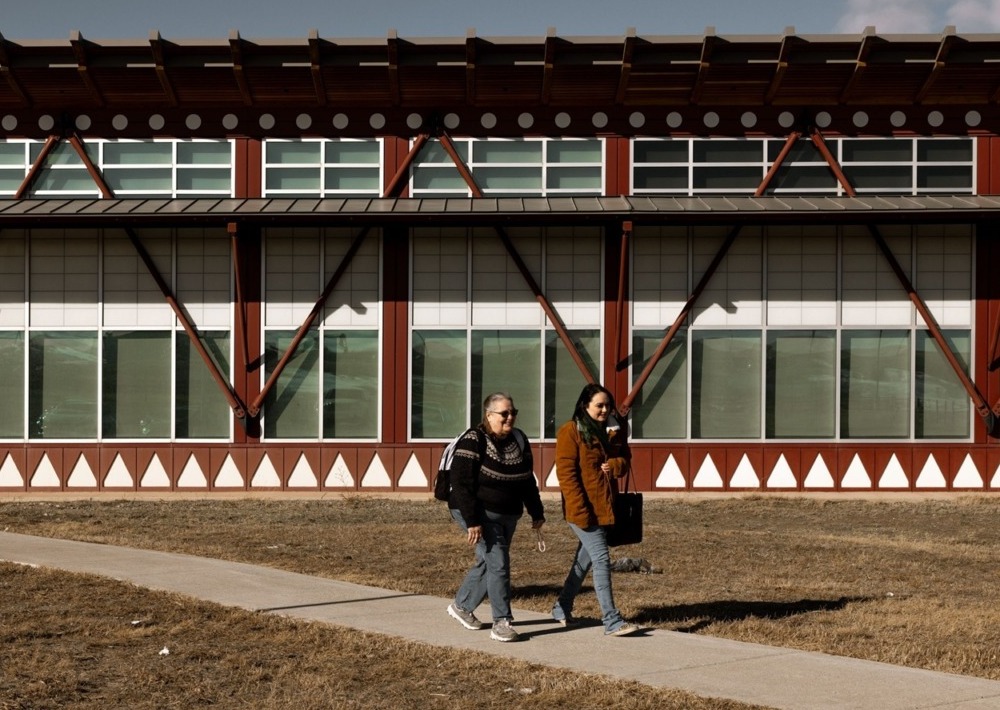 Trump wants to cut tribal college funding by nearly 90%, putting them at risk of closing: two people walk on sidewalk away from campus building on nice day