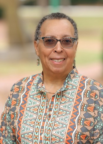 Karen Pittman opinion: headshot black woman with very short hair, dark-framed glasses wearing a colorful pattern top in front of outdoor trail with greenry