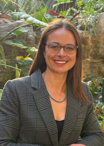 Katherine Plog Martinez: white woman with long, dark curly hair, black rim glasses, wearing purple jacket, smiling outside in front of trees