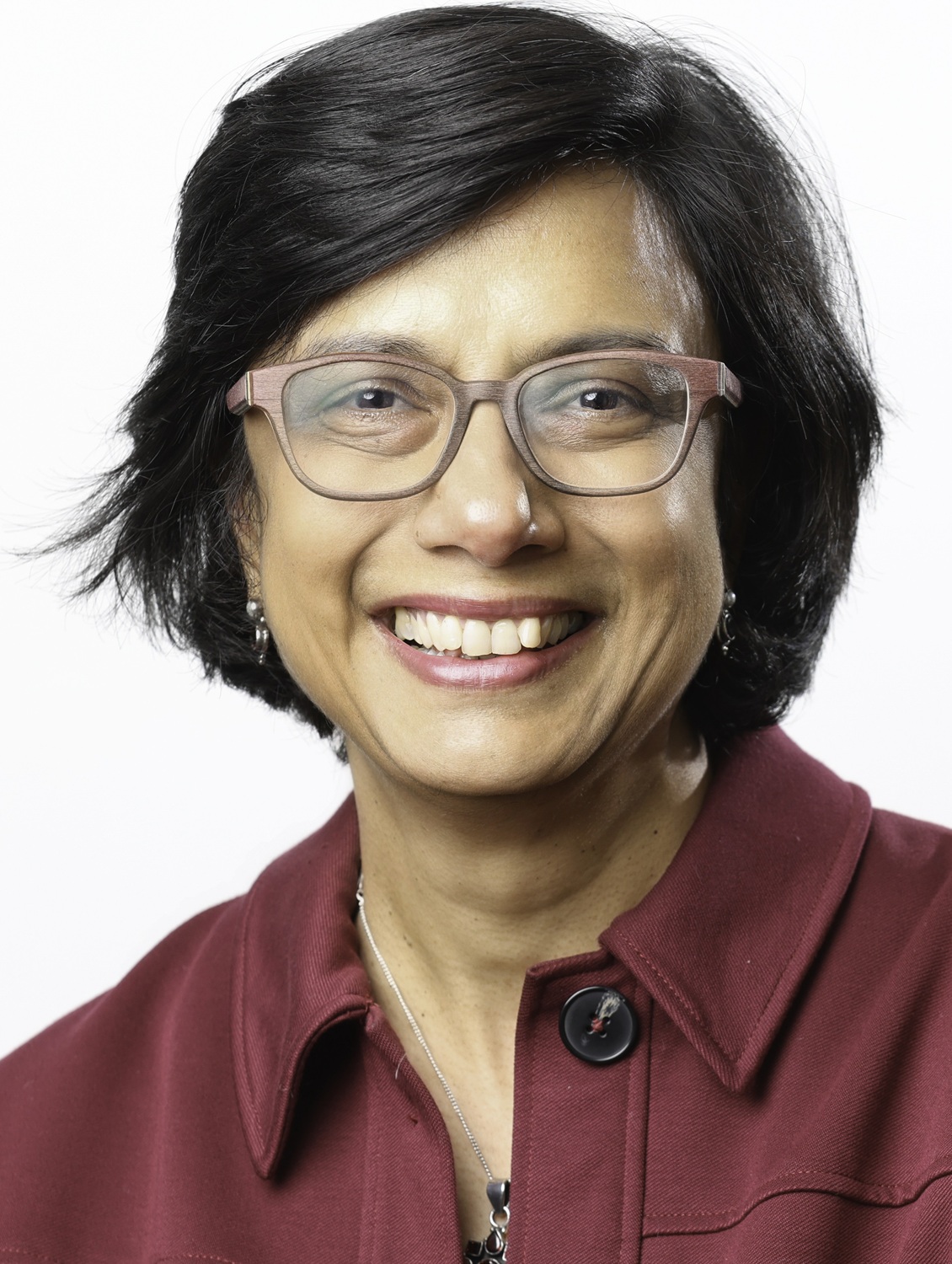 Anita Krishnamurthi headshot: woman with short dark hair and thick-rimmed glasses smiles in front of white background