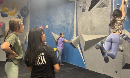 From athlete to youth sports coach_A new ascent_feature: two young women watch on as another swings legs out from climbing wall