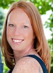 Heather Kennedy headshot: white woman with red hair outdoors smiling over shoulder