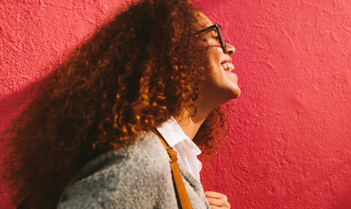 foster youth; Never too young to be an advocate_feature: Side view of smiling young woman with curly hair against red wall. Laughing african young woman with bag.
