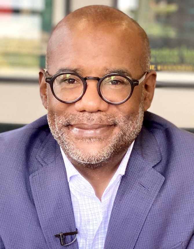 Bryan Samuels headshot: black, bald man in round-rimmed glasses in suit jacket at desk