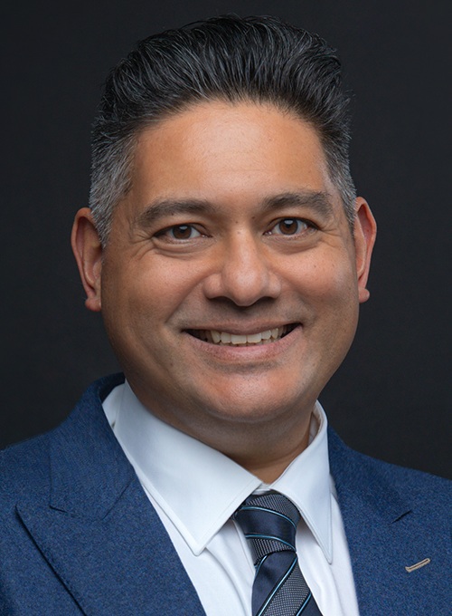 صورة لورانس غانتي في الرأس: رجل يرتدي بدلة زرقاء داكنة وربطة عنق أمام خلفية رمادية داكنة