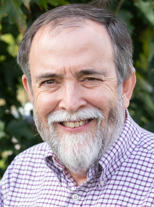Gene Roehlkepartain headshot: older white man with grey hair and white beard smiling outdoors wearing collared patterned shirt
