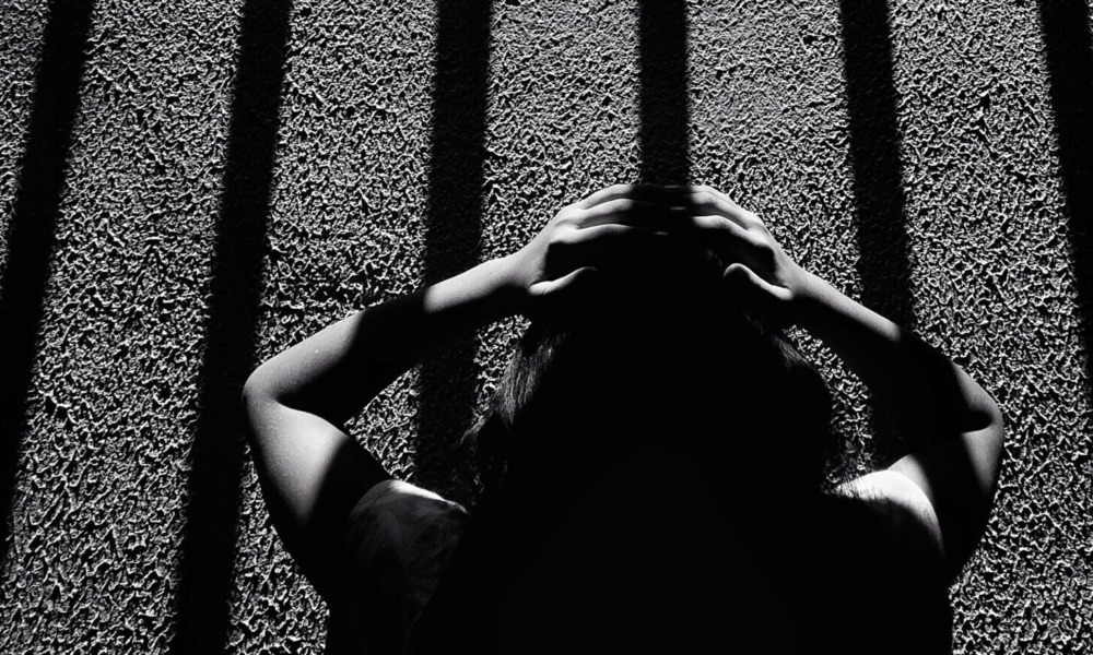 Twice a refugee _feature: This black-and-white image depicts a dramatic scene of a person holding their head, seemingly distressed, while standing under long, vertical shadows resembling prison bars. The composition and the strong contrast between light and dark create an intense atmosphere, evoking feelings of confinement, mental struggle, and isolation. The texture of the ground and the person's silhouette add depth to the emotional narrative of the photograph.