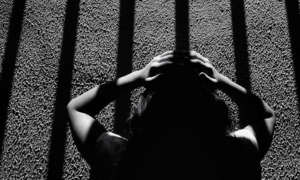 Twice a refugee _feature: This black-and-white image depicts a dramatic scene of a person holding their head, seemingly distressed, while standing under long, vertical shadows resembling prison bars. The composition and the strong contrast between light and dark create an intense atmosphere, evoking feelings of confinement, mental struggle, and isolation. The texture of the ground and the person's silhouette add depth to the emotional narrative of the photograph.