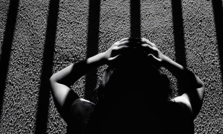 Twice a refugee _feature: This black-and-white image depicts a dramatic scene of a person holding their head, seemingly distressed, while standing under long, vertical shadows resembling prison bars. The composition and the strong contrast between light and dark create an intense atmosphere, evoking feelings of confinement, mental struggle, and isolation. The texture of the ground and the person's silhouette add depth to the emotional narrative of the photograph.