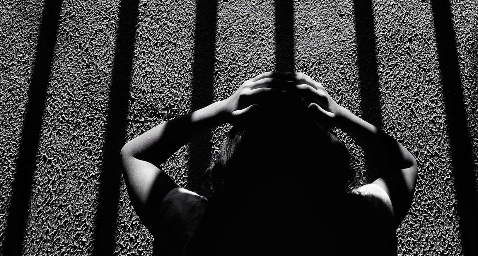 Twice a refugee _feature: This black-and-white image depicts a dramatic scene of a person holding their head, seemingly distressed, while standing under long, vertical shadows resembling prison bars. The composition and the strong contrast between light and dark create an intense atmosphere, evoking feelings of confinement, mental struggle, and isolation. The texture of the ground and the person's silhouette add depth to the emotional narrative of the photograph.