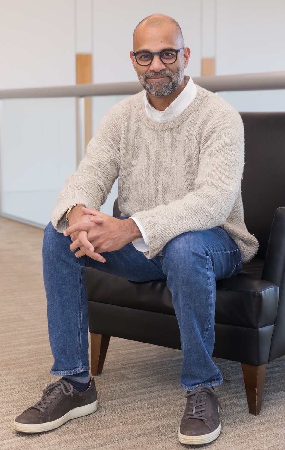 Rajiv Vinnakota headshot2: bald man in thick-rimmed glasses with greying short beard sitting in chair with hands clasped