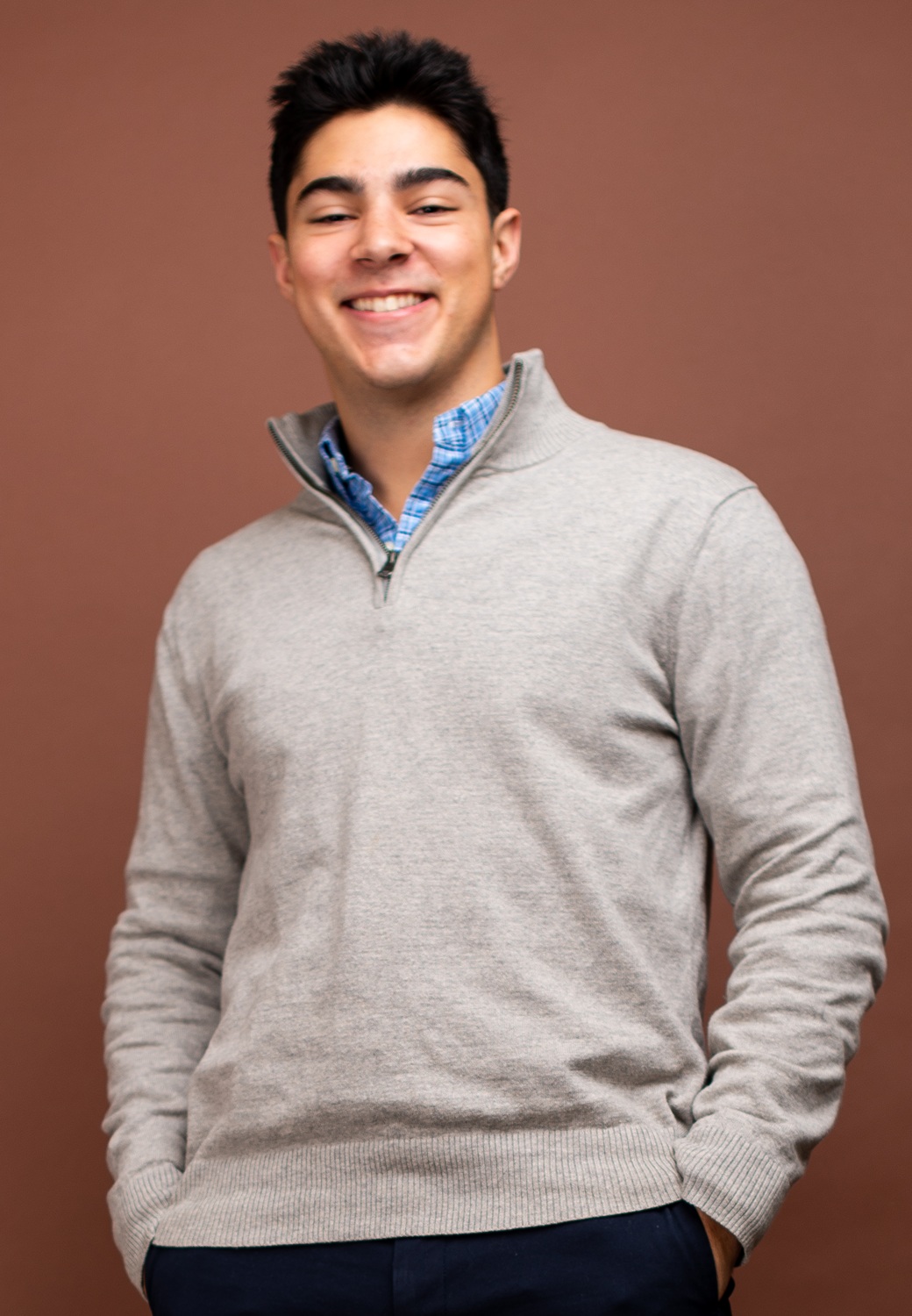 Sam Brill headshot: young man with black hair wearing grey sweater in front of red background