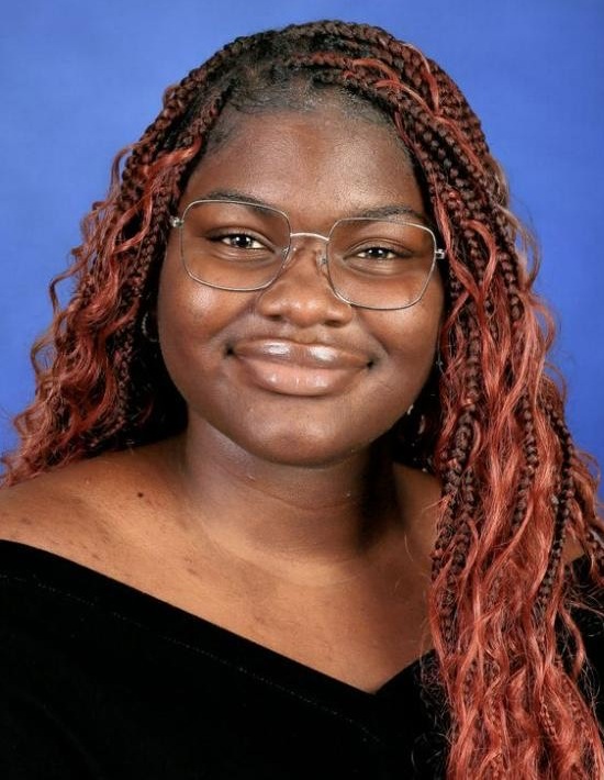 D'Avora Williams headshot: young black woman with long curly hair wearing black in front of blue background