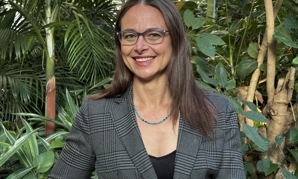 Youth Today Katherine Plog Martinez headshot_feature: woman with long brown hair and glasses smiling in front of plants