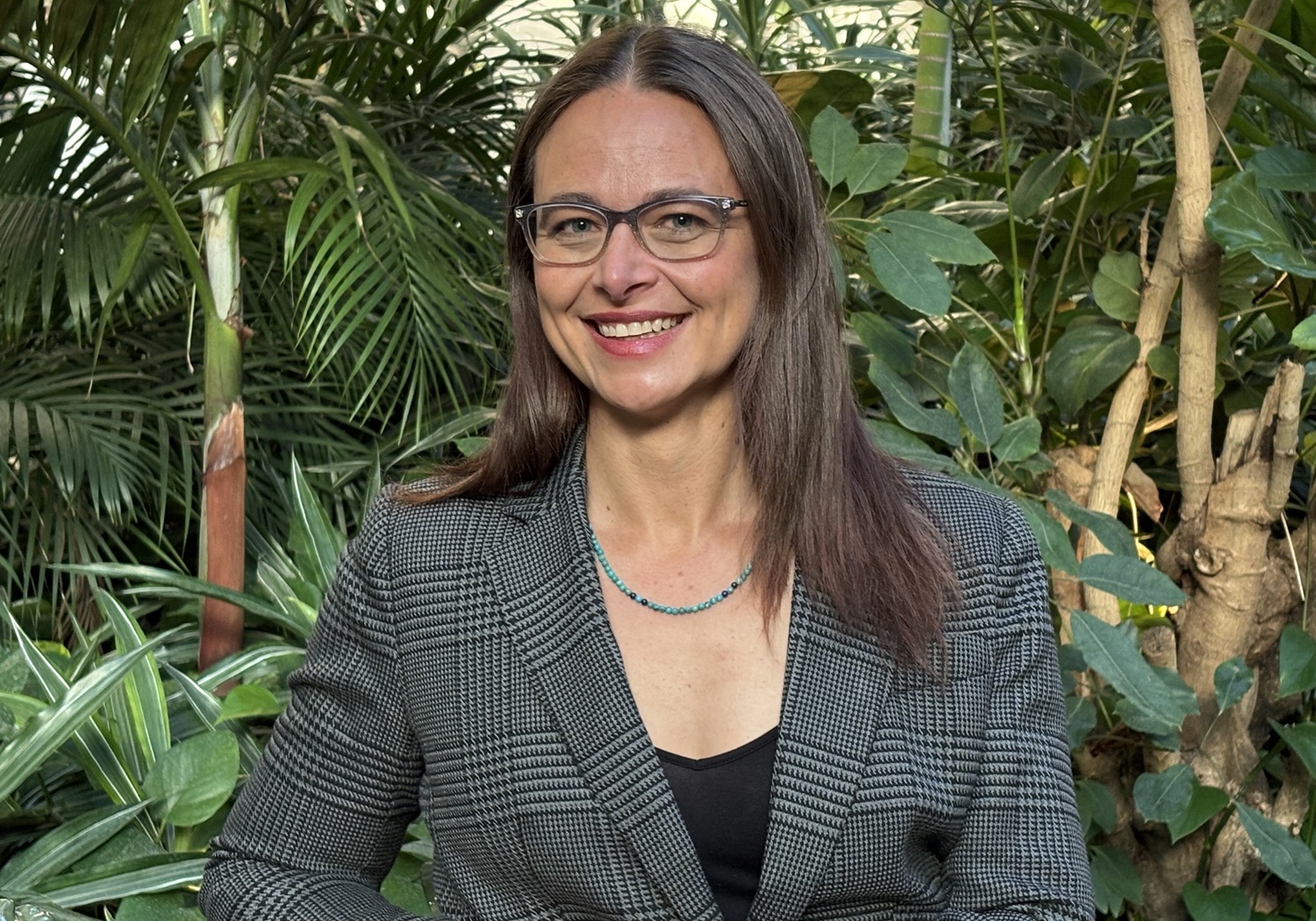 Youth Today Katherine Plog Martinez headshot_feature: woman with long brown hair and glasses smiling in front of plants