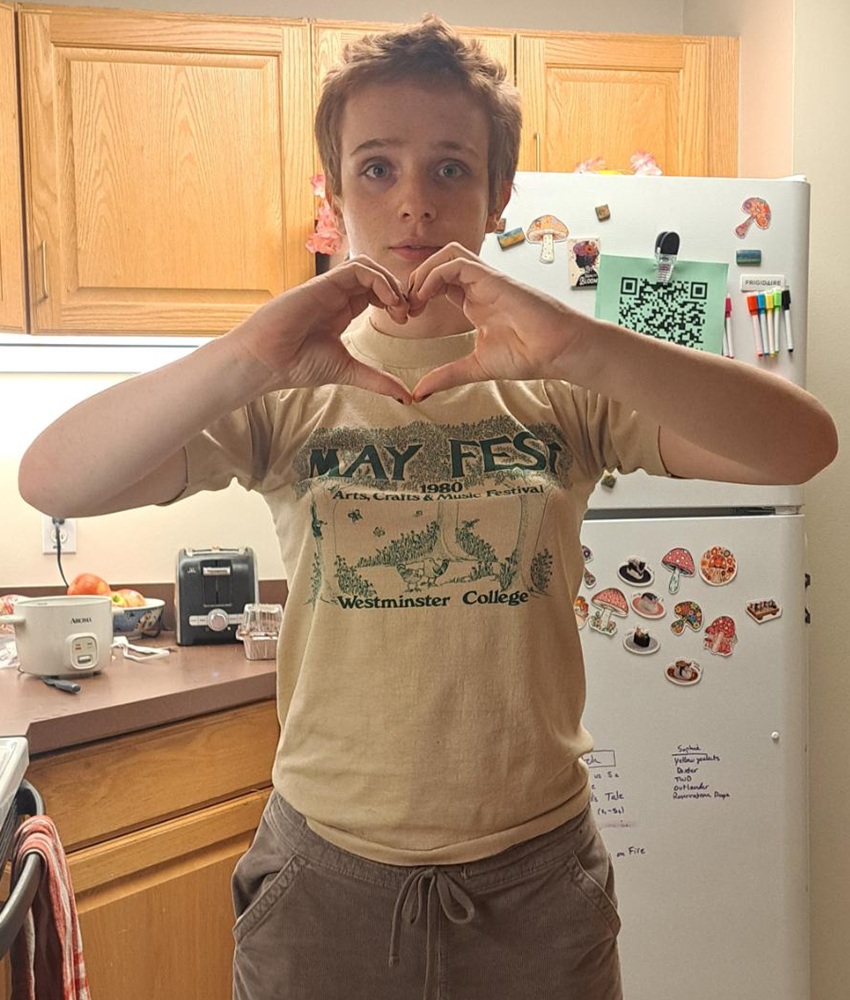 Harper student headshot: young student with short hair standing in kitchen making heart shape with hands
