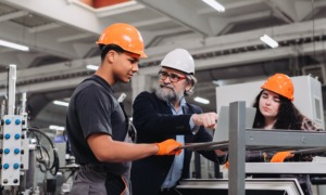 Skilled trades training and capacity-building program grants: older tradesman teaches young male and female in training on factory floor