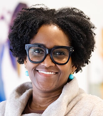 Audra Watson headshot: black woman with black hari and thick rimmed glasses in front of colorful background