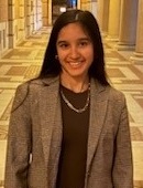 Deepika Miryala headshot: young woman with long dark hair standing in hallway wearing shirt and blazer with necklace