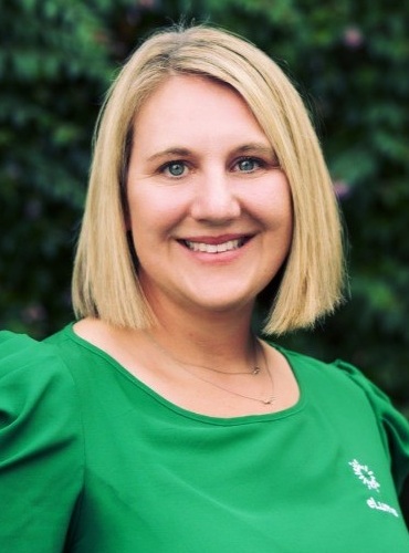 Kay Kelly headshot: white woman with medium length blonde hair smiling and wearing green shirt outdoors