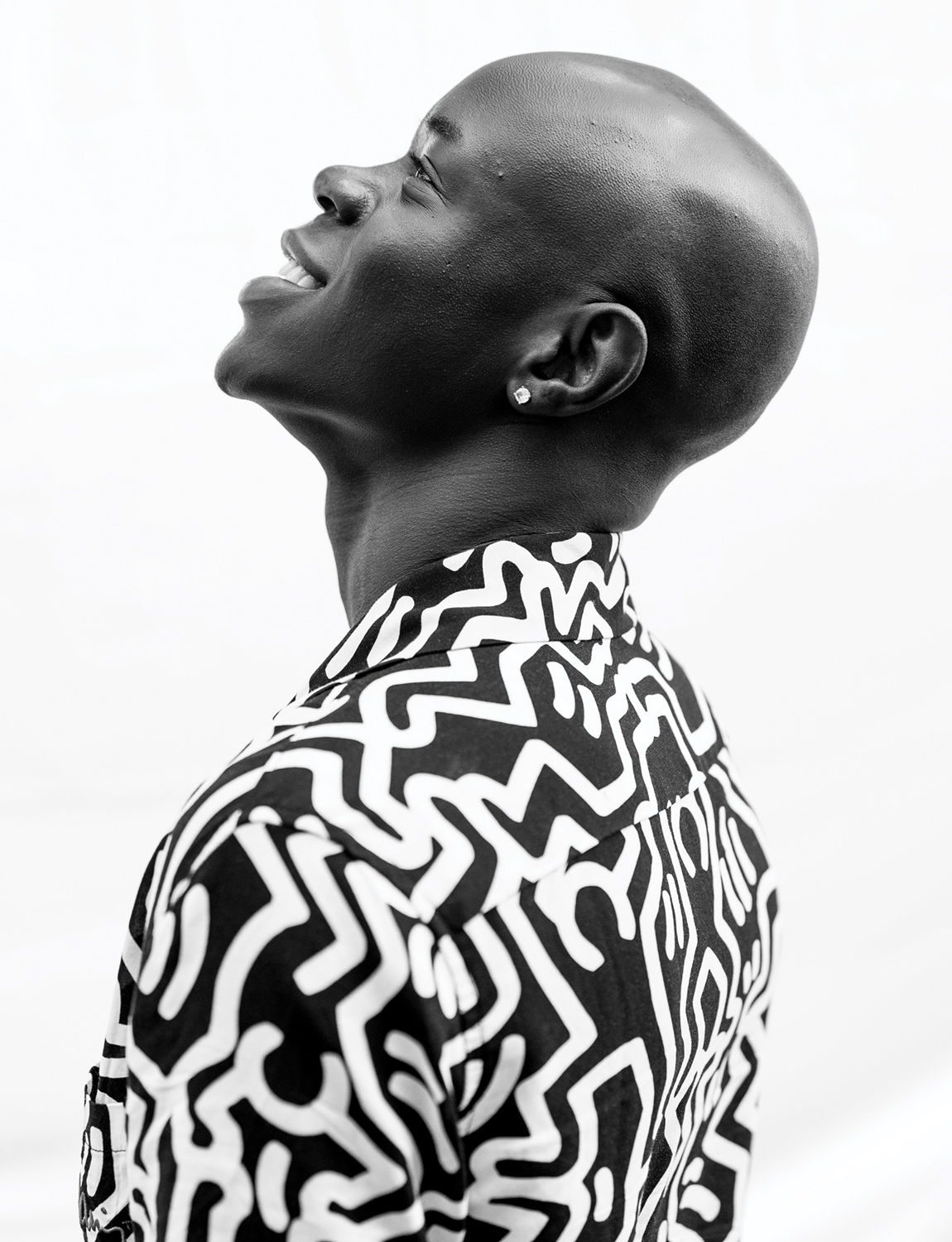 Gabriel Arika headshot: side view of bald black man smiling and looking upwards while wearing black and white shirt