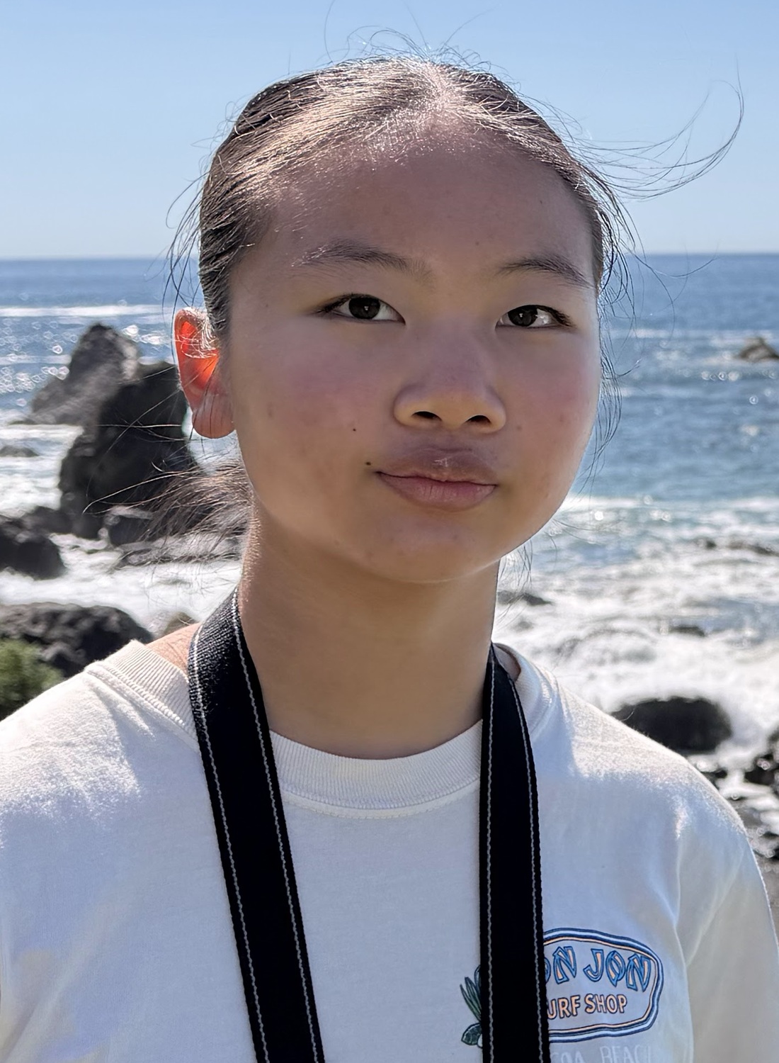 Gloria Chi headshot: young Asian woman in white t-shirt and lanyard around neck standing at seashore with ocean waves breaking on rocks in the background