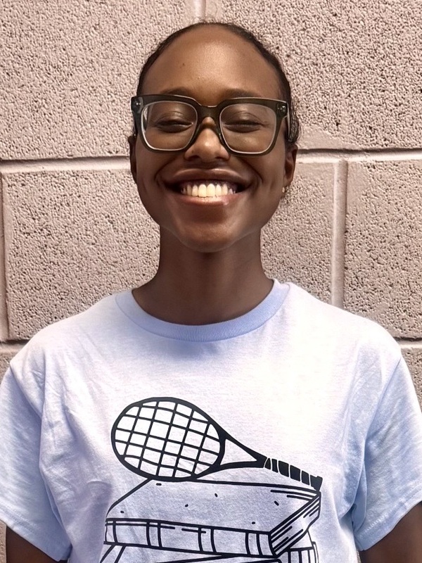 Deja Martyr headshot: young black teen wearing glasses and tennis shirt standing in front of cinder block wall