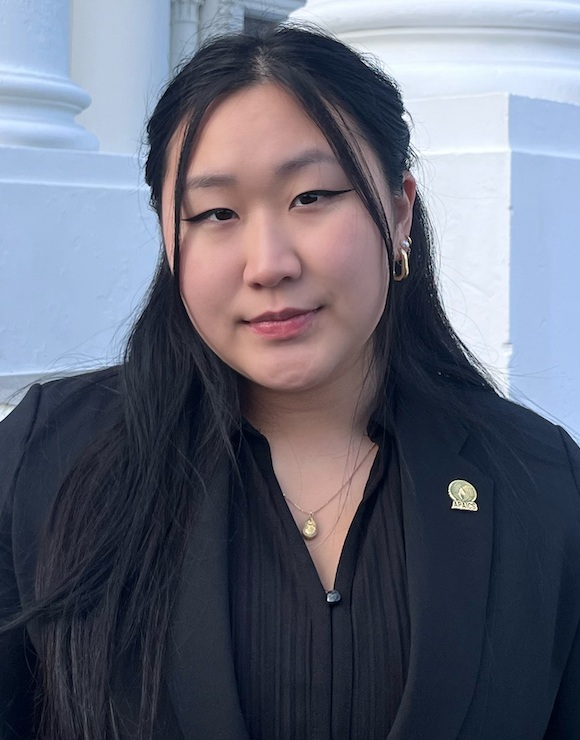 Maddy Park headshot: Asian woman with long black hair wearing black blazer and shirt in front of white building